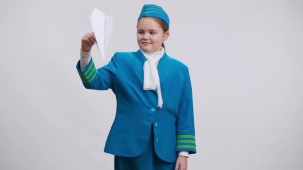 Söt flicka i flygvärdinna uniform spelar landning papper plan vänder sig till kameran leende. Porträtt av charmiga vita barn drömmer om flygvärdinna jobb poserar på vit bakgrund. — Stockvideo