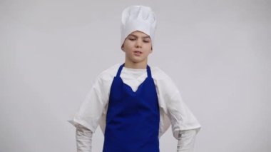 Stresli küçük aşçı şakakları ovuyor, şef şapkasını kafasına geçiriyor. Beyaz arka planda poz veren yorgun beyaz çocuk portresi. Mutfak ve mutfak mesleği seçimi.