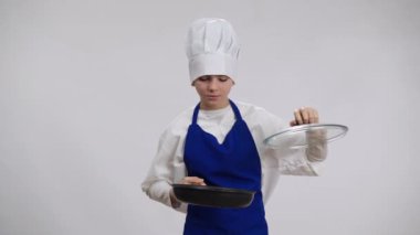 Kendinden emin küçük aşçı kızartma tavasını açıyor lezzetli kokulu yemekler yiyor gururlu bir yüz ifadesiyle kameraya bakıyor. Şef şapkalı ve önlüklü, arka planda poz veren tatmin olmuş beyaz çocuk portresi..
