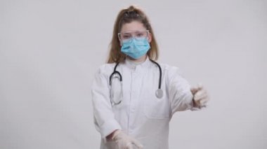 Coronavirus 'lu küçük hemşire maske takıyor, gözlük takıyor ve beyaz arka planda el ele tutuşuyorlar. Tıp mesleğini seçen ciddi bir beyaz kızın portresi kameraya bakıyor..