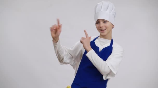 Χαρούμενο χαρούμενο αγόρι με καπέλο μάγειρα και ποδιά να δείχνει στην άκρη κοιτάζοντας την κάμερα με χαμόγελο στα δόντια. Πορτρέτο του χαρούμενου καυκάσιου παιδιού που ποζάρει σε λευκό φόντο επιλέγοντας επάγγελμα μαγειρικής. — Αρχείο Βίντεο
