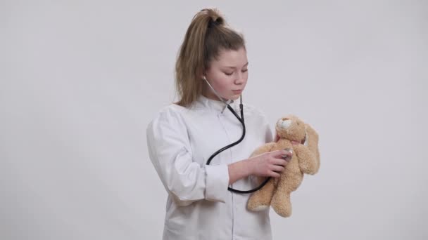 Concentrated Caucasian girl in doctor gown listening teddy bear with stethoscope and looking at camera. Portrait of confident focused child choosing medical profession posing at white background. — Stock Video