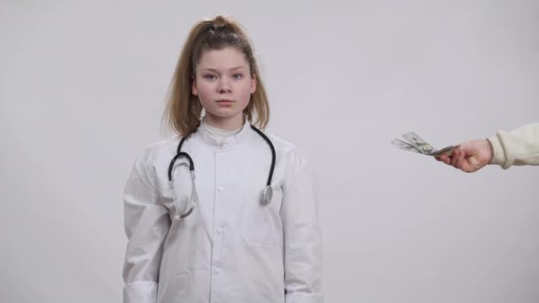 Porträtt av en liten läkare som vägrar muta en vit bakgrund. Självsäker ärlig vit flicka väljer läkaryrket avvisa pengar tittar på kameran. Begreppet korruption inom hälso- och sjukvård. — Stockvideo