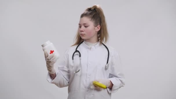 Καυκάσιος γιατρός διαλέγει μεταξύ χαπιών και μπανάνας που απλώνει φρούτα στην κάμερα κουνώντας το κεφάλι ναι. Πορτρέτο του κοριτσιού με αυτοπεποίθηση ποζάρουν με φάρμακα και οργανική επιλογή βιταμινών σε λευκό φόντο — Αρχείο Βίντεο