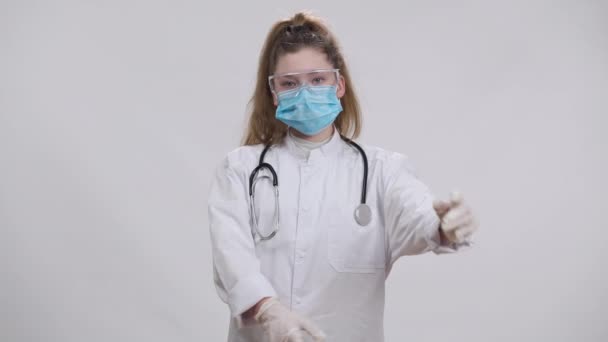 Little nurse doctor in coronavirus face mask medical gown and eyeglasses crossing hands posing at white background. Portrait of serious Caucasian girl choosing medicine profession looking at camera. — Stock Video