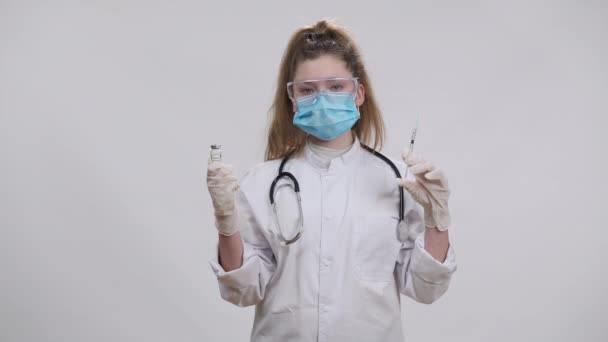 Уверенный маленький доктор в маске для лица Ковида держит вакцину Ковид-19, джеб и шприц, смотрящий в камеру с серьезным выражением лица. Портрет белой девушки, позирующей с пандемическими лекарствами. — стоковое видео