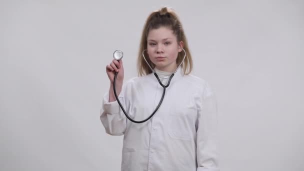 Портрет уверенной девушки, показывающей стетоскоп, смотрящей в камеру, улыбающейся стоя на белом фоне в халате врача. Очаровательный кавказский ребенок выбирает медицинскую профессию. — стоковое видео