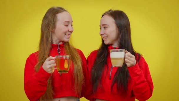 Slim charming twin sisters clinking cups with tea and coffee drinking smiling looking at camera. Portrait of happy Caucasian beautiful women posing at yellow background with hot drink. — Stock Video