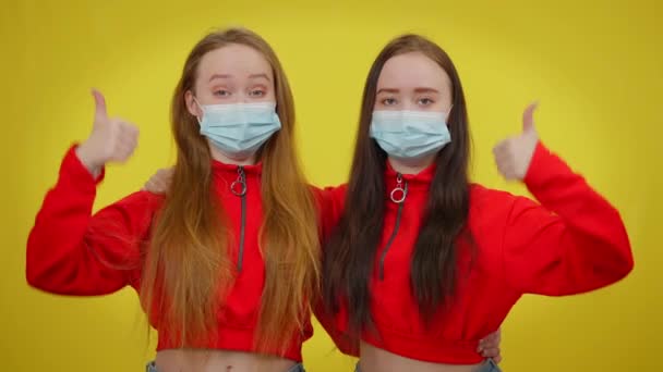 Young twins in coronavirus face masks gesturing thumb up looking at camera. Portrait of confident Caucasian beautiful slim women posing at yellow background on Covid-19 pandemic. — Stock Video