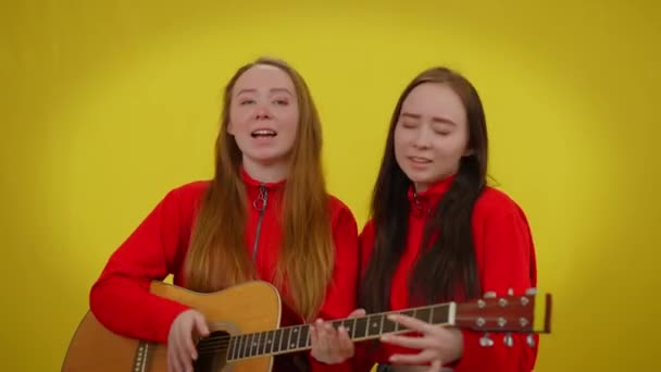 Gemelos felices cantando la guitarra en el fondo amarillo sonriendo. Retrato de hermosas mujeres jóvenes caucásicas disfrutando de hobby estilo de vida escribiendo canción. Concepto de felicidad y unidad. — Vídeo de stock