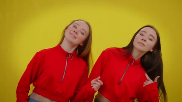 Giovani donne sorridenti che si muovono simultaneamente ballando su sfondo giallo guardando la fotocamera. Fiduciose sorelle gemelle caucasiche in posa godendo della musica. — Video Stock