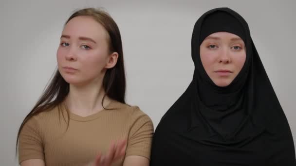 Självsäker smal kvinna i hijab tittar på kameran som tvillingsyster korsar händer gestikulerande nej. Unga kaukasiska kvinnliga tvillingar poserar på grå bakgrund. Begreppet religion och individualitet. — Stockvideo