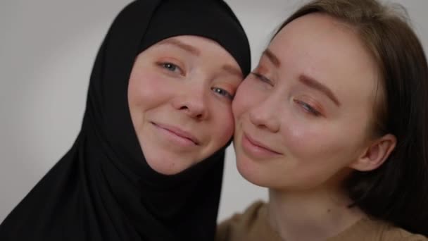 Visi ravvicinati di sorelle gemelle che guardano una telecamera che sorride posando su uno sfondo grigio. Giovane donna caucasica positiva e fratello in hijab nero faccia a faccia. Differenza e individualità. — Video Stock