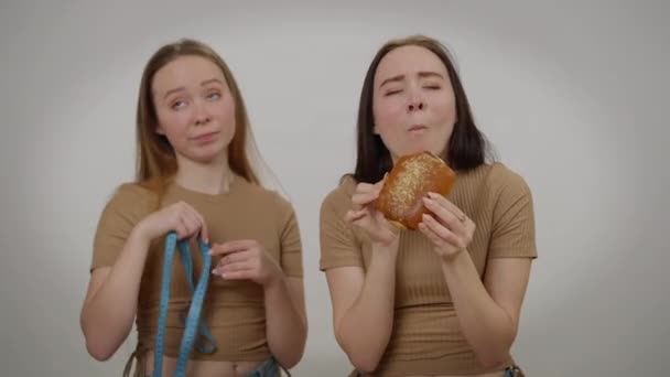 Молодая женщина наслаждается вкусом нездоровой сладкой булочки, как сестра-близнец показывает измерительную ленту. Идентичные белые братья и сёстры на сером фоне. Концепция самоконтроля и диеты. — стоковое видео