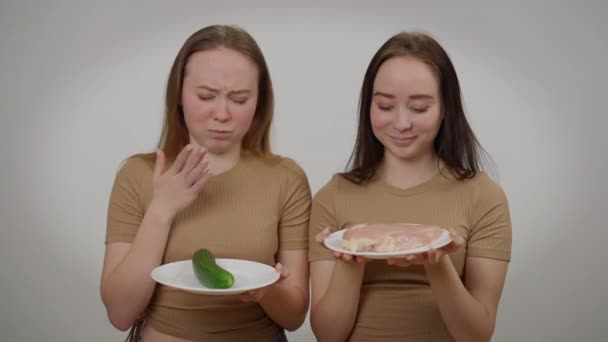 Tweeling zussen poseren met rauw vlees en verse biologische komkommer op grijze achtergrond. Portret van zelfverzekerde vleeseter en veganistische blanke vrouwen die glimlachend naar de camera kijken. — Stockvideo