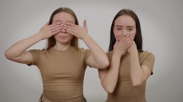 Az ikrek becsukják a szemüket és a szájukat, és komoly arckifejezéssel néznek a kamerába. Közepes felvételű portré magabiztos kaukázusi nőkről, akik szürke hátterű gesztikulálva pózolnak. — Stock videók