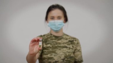 Bulanık yüzlü kadın asker Coronavirus aşısı pozu veriyor gri arka planda. Kendinden emin, ciddi, beyaz, genç bir kadının pandemik tedaviyi elinde tutması..