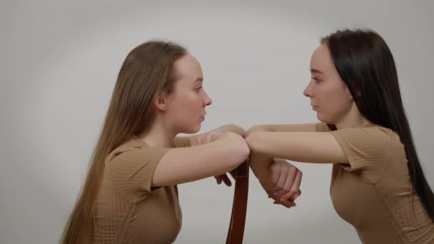 Des jeunes sœurs jumelles concentrées se regardant se tournant vers la caméra et le dos. Vue de côté portrait de graves belles femmes caucasiennes assises sur des chaises posant sur fond gris. — Video