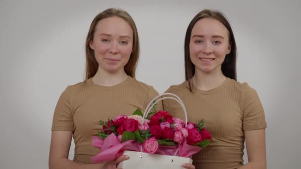 Dois sorrindo belas mulheres idênticas posando com cesta de flores em fundo cinza. Retrato de confiantes encantadoras irmãs gêmeas caucasianas olhando para a câmera com buquê. Femininidade e irmandade. — Vídeo de Stock