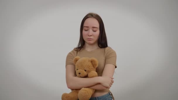 Sfrustrowana młoda kobieta stojąca z misiaczkiem patrząca w kamerę ze smutnym wyrazem twarzy. Portret zdenerwowanej pięknej szczupłej białej kobiety pozowanie na szarym tle przytulanie zabawki. — Wideo stockowe