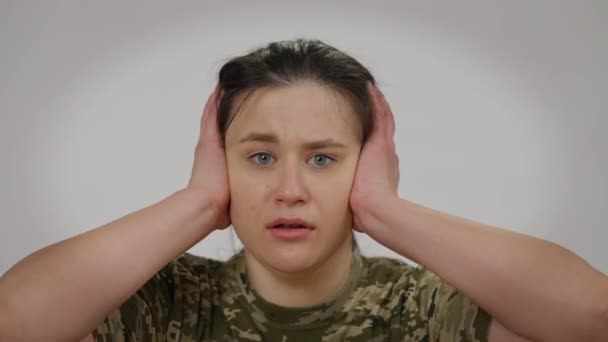 Retrato de cerca de una aterrorizada militar mirando a la cámara cerrando las orejas con las manos. Joven y asustada hermosa soldado caucásica posando en un fondo gris. Concepto de pánico y miedo. — Vídeos de Stock