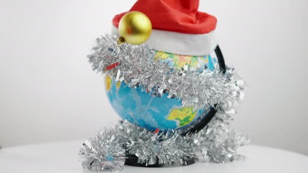 Close-up globo terrestre em chapéu de Natal e guirlanda decorativa com bolas de Ano Novo caindo na mesa em câmera lenta. Conceito de feriados temporada celebração global. Tradições e festivais. — Vídeo de Stock