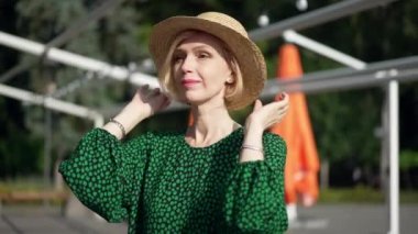 Açık havada güneşli bir günde yeşil elbiseli ve hasır şapkalı zayıf, kendine güvenen yetişkin bir kadının görüntüsü. Kafkasyalı mutlu, rahat bir bayanın hafta sonunun tadını çıkaran portresi. Yavaş çekim.