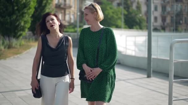 Dos mujeres adultas seguras caminando en la ciudad hablando en cámara lenta riendo. Retrato de vista frontal Mujeres amigas caucásicas paseando al aire libre en un día soleado chismorreando sonriendo. Amistad y estilo de vida. — Vídeo de stock