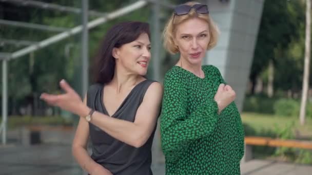 Dos delgadas mujeres caucásicas adultas haciendo gestos de fuerza riendo de espaldas al aire libre. Amigos felices charlando posando en la calle de la ciudad. Confianza y estilo de vida. — Vídeos de Stock