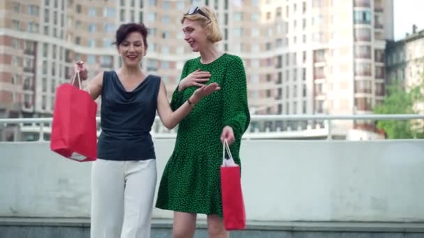 Dos mujeres felices caminando por la ciudad con bolsas de compras sonriendo y hablando en cámara lenta. Adultas amigas caucásicas disfrutando de la temporada de ventas del Viernes Negro al aire libre. Consumismo y estilo de vida. — Vídeo de stock