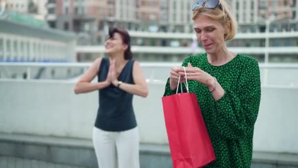 Mujer adulta satisfecha regocijándose Viernes Negro abrazando bolsa de compras con alegre amigo sonriendo de fondo. Retrato de caucásico feliz shopaholic al aire libre en la calle de la ciudad. Movimiento lento. — Vídeo de stock
