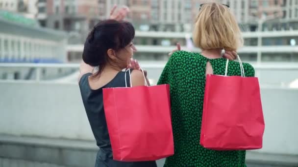 バックビューショッピングバッグと2人の興奮した女性は、屋外でブラックフライデーを楽しんで笑顔踊ります。中規模の幸せな白人の買い物客が街の路上で話して立って撮影した。販売と友情. — ストック動画