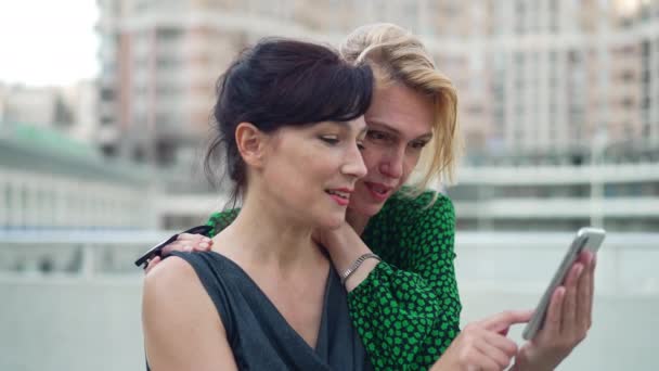 Πλευρική άποψη χαρούμενη ενήλικες γυναίκες κύλιση social media στο smartphone μιλώντας χαμογελώντας σε αργή κίνηση. Πορτρέτο των χαρούμενων Καυκάσιων φίλους surfing Internet απολαμβάνοντας Σαββατοκύριακο στο δρόμο της πόλης. — Αρχείο Βίντεο