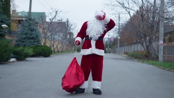 Široký záběr Santa Clause chůze na předměstské ulici s bolestí zad zavěšení dárkové tašky na rameni odchodu. Portrét bělocha v červeném kostýmu na Štědrý večer venku procházky pryč. — Stock video