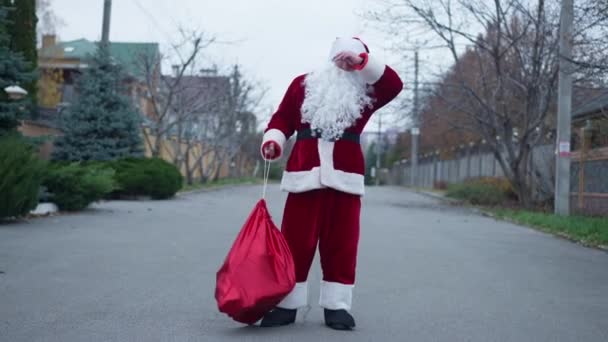 Grande colpo Babbo Natale Clause a piedi sulla strada suburbana avendo mal di schiena appeso sacchetto regalo sulla partenza a spalla. Ritratto di uomo caucasico in costume rosso la vigilia di Natale all'aperto passeggiando via. — Video Stock