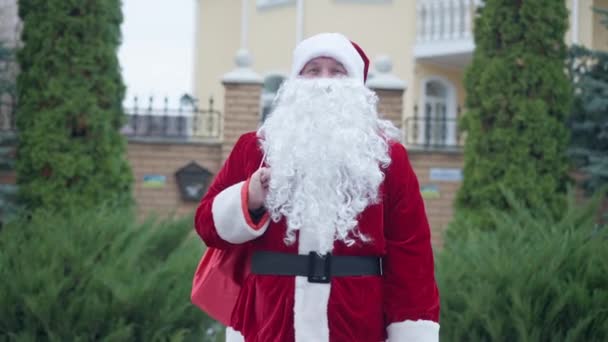 Porträtt av vit man i röd jultomtedräkt med skägg viftande tittar på kameran leende. Medium skott positiv ung kille med presentpåse poserar utomhus på semester. Nytt år och jul. — Stockvideo