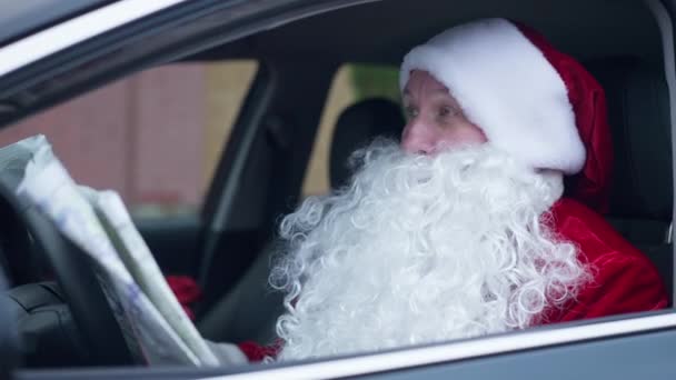 Förvirrad skäggig jultomte sittande i bil på förarsätet undersöker papperskarta. Porträtt av förlorade jultomten i fordonet utomhus söker rätt väg att gå. Nyårsresor. — Stockvideo