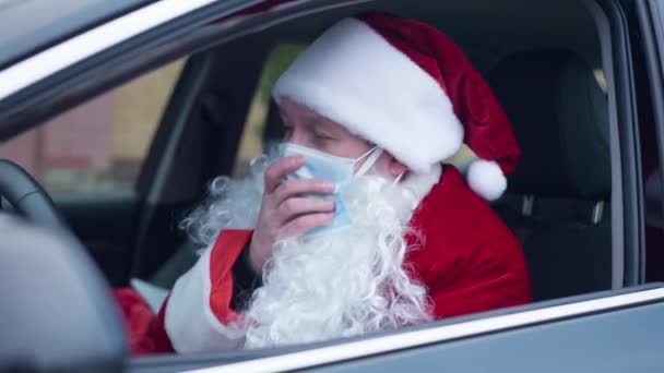 Ik zal de kerstman in coronavirus masker hoesten zittend in de auto buiten. Trieste onwel Kaukasische man in rood kostuum met baard werkt aan Covid-19 pandemische kerstvakantie. — Stockvideo