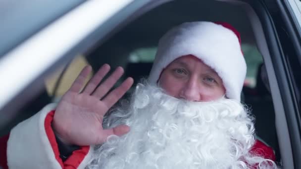 Santa Clause agitant en regardant la caméra assise sur le siège du conducteur dans la voiture. Portrait du jeune Père Noël caucasien moderne posant dans un véhicule à l'extérieur la veille du Nouvel An. Fête célébration. — Video