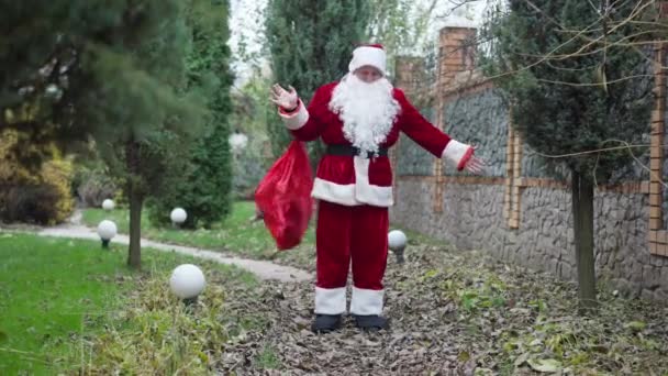 Široký záběr Santa s dárky taška stojí na dvorku se zelenými stromy. Portrét mladého bělošského pozitivního muže v kostýmu s vousy a představuje venku. Koncept Vánoc a Nového roku. — Stock video