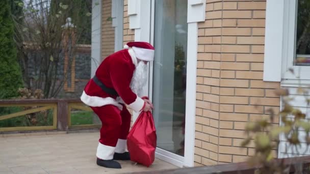 Szerokie ujęcie złodzieja w kostiumie Świętego Mikołaja otwierającego drzwi na podwórku w Sylwestra. Młody biały mężczyzna włamywacz okrada dom w święta. Łamacz złodziei rozgląda się po otwartych drzwiach. — Wideo stockowe