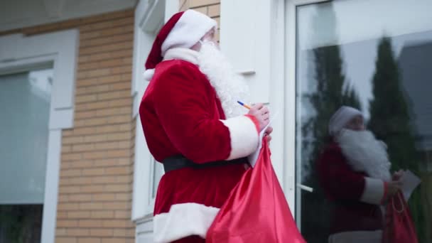 Ung man i tomtedräkt skriver på brev kuvert gå iväg med presentpåse. Positivt självsäker vit jultomte utomhus på bakgården lämnar. Nyårsfirande och överraskningskoncept. — Stockvideo