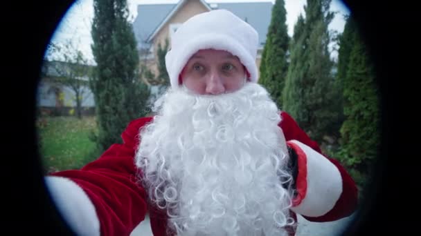 Utsikt genom titthål Santa Clause ringer dörrklocka stående på grön trädgård utomhus. Front view man i röd jultomte med vitt skägg och presentpåse kommer på nyår. — Stockvideo