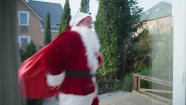 Αστείος Άγιος Βασίλης τρέχει με τα πόδια με τα δώρα τσάντα στην αυλή σε εξωτερικούς χώρους. Σκοποβολή μέσα από το γυάλινο παράθυρο του χαρούμενου Καυκάσιου άνδρα στο κοστούμι του Πατέρα Χριστούγεννα διασκεδάζοντας. — Αρχείο Βίντεο