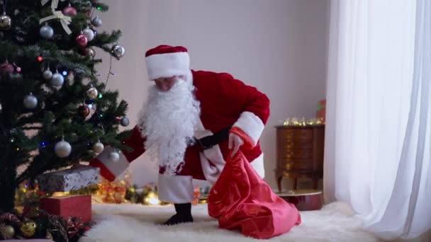 Jultomten lägger inslagna julklappar under julgranen i vardagsrummet inomhus. Brett skott positiv vit man i röd kostym med gåvor. Nyår mirakel och överraskning koncept. — Stockvideo