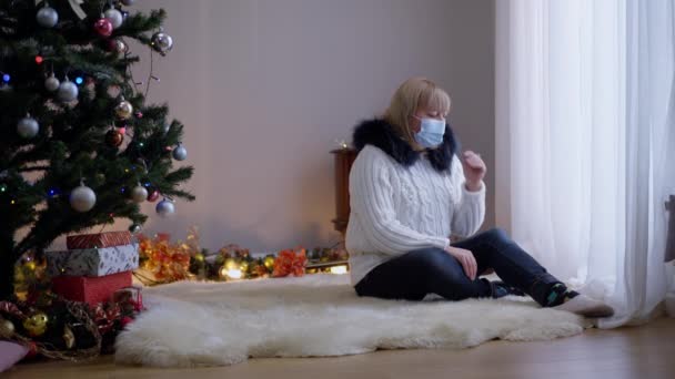 Sorglig kvinna i coronavirus ansikte mask gråter på julen ensam hemma som jultomten in i vardagsrummet ger gåva. Brett skott deprimerad kaukasisk sjuk dam får närvarande på Covid-19 nyår. — Stockvideo