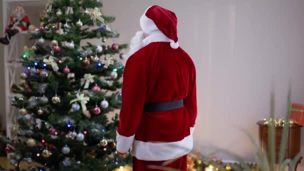 Dumny biały Mikołaj podziwiający ozdobioną choinkę w salonie. Średni strzał pewny siebie człowiek w Ojciec Boże Narodzenie kostium dekorowanie domu w Sylwestra w pomieszczeniach. — Wideo stockowe