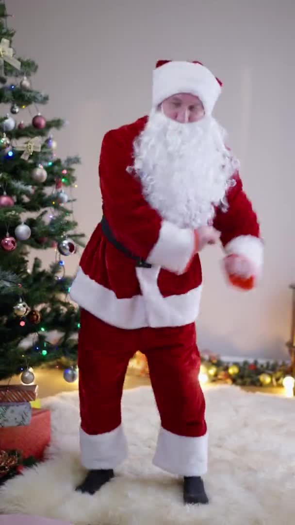 Vertikal video glad jultomte dans inomhus i vardagsrummet på dekorerade nyårsträd. Brett skott glad glad glad vit jultomte jultomten ha kul på semester kväll. — Stockvideo
