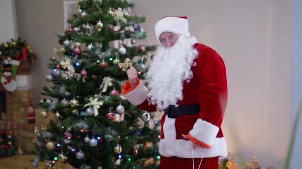 Портрет радостного Санты, указывающего на украшенную новогоднюю елку с жестом молчания пальцем на губах. Белый молодой человек в красном костюме Деда Мороза в гостиной. Сюрприз. — стоковое видео