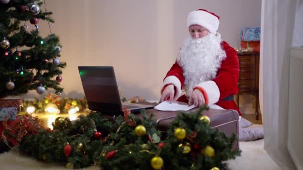 Osäker upptagen jultomten läsa bokstäver trycka på tangenterna på laptop inomhus. Porträtt av generad vit jultomte som handlar om modern teknik på semester kväll i vardagsrummet. — Stockvideo